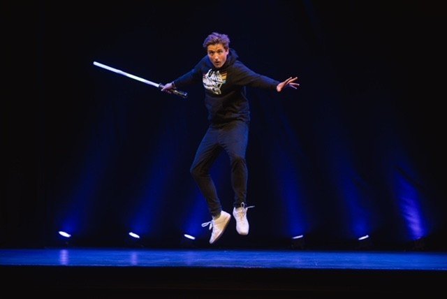 This image shows a person captured mid-air on a stage, appearing to perform or pose dynamically while holding a glowing, sword-like prop.
