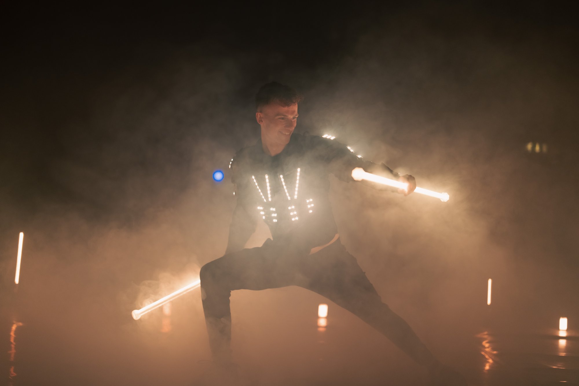 A performer in an illuminated LED costume wields glowing light batons amid theatrical haze, creating a striking light and movement spectacle.