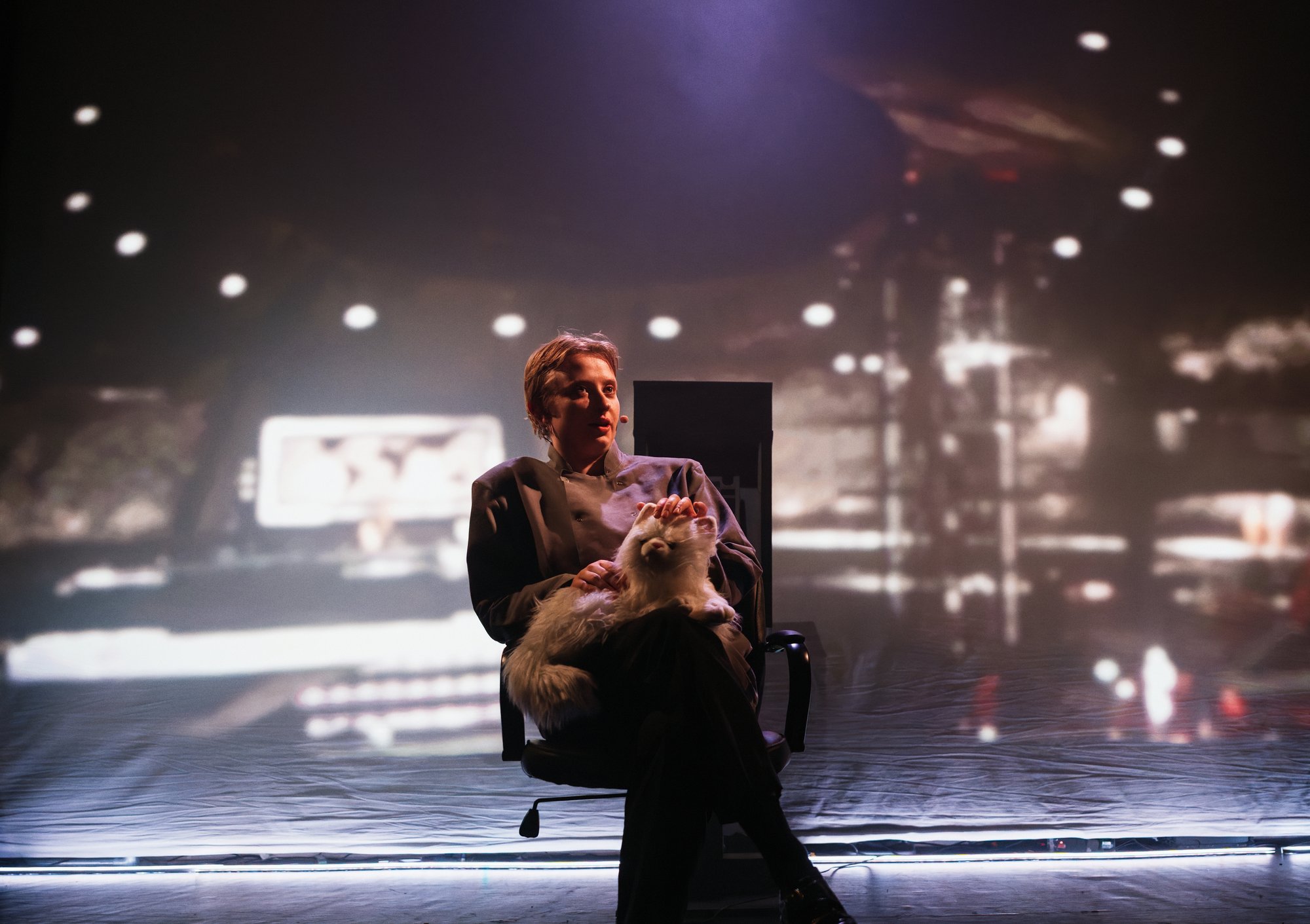 A single person is seated in a black office-style chair at centre stage, while a large projected image or backdrop fills the background.