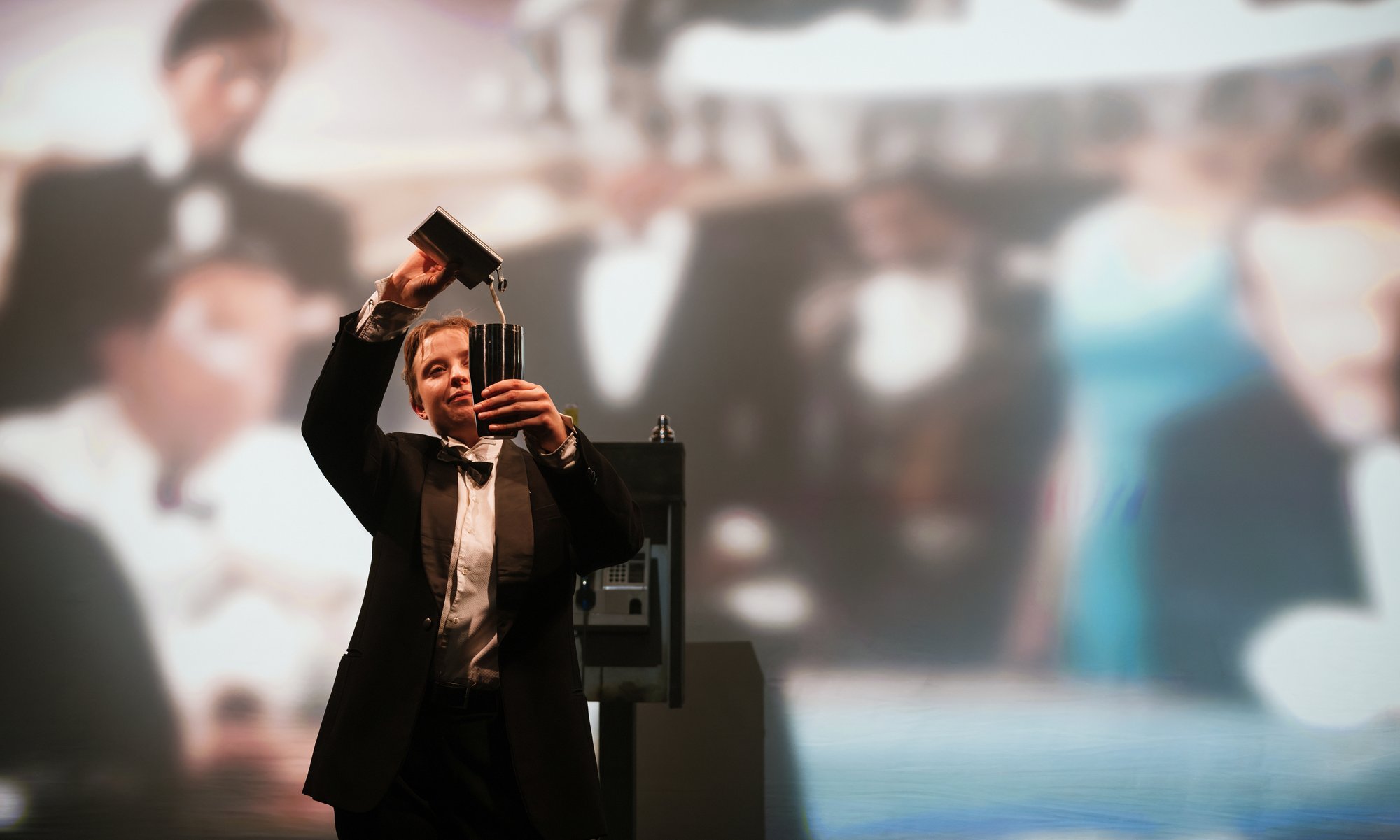 In the foreground, a person wearing formal evening attire, including a black tuxedo-style jacket, white shirt, and black bow tie, is holding a tall metal cocktail shaker or mixing vessel.