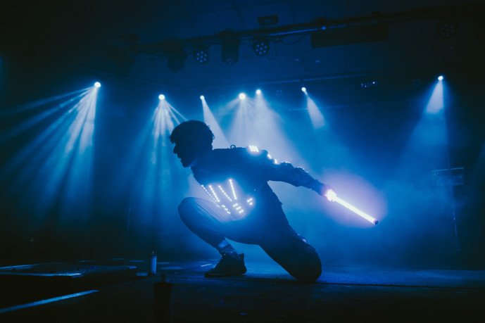 A performer in an illuminated LED costume on a dark stage, holding a glowing light wand, surrounded by blue stage lighting and beams of light.