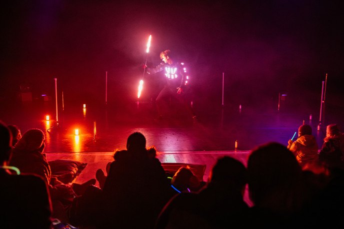 An LED-lit performer stands at the centre of a stage, spinning glowing light staffs amid red and pink lighting effects