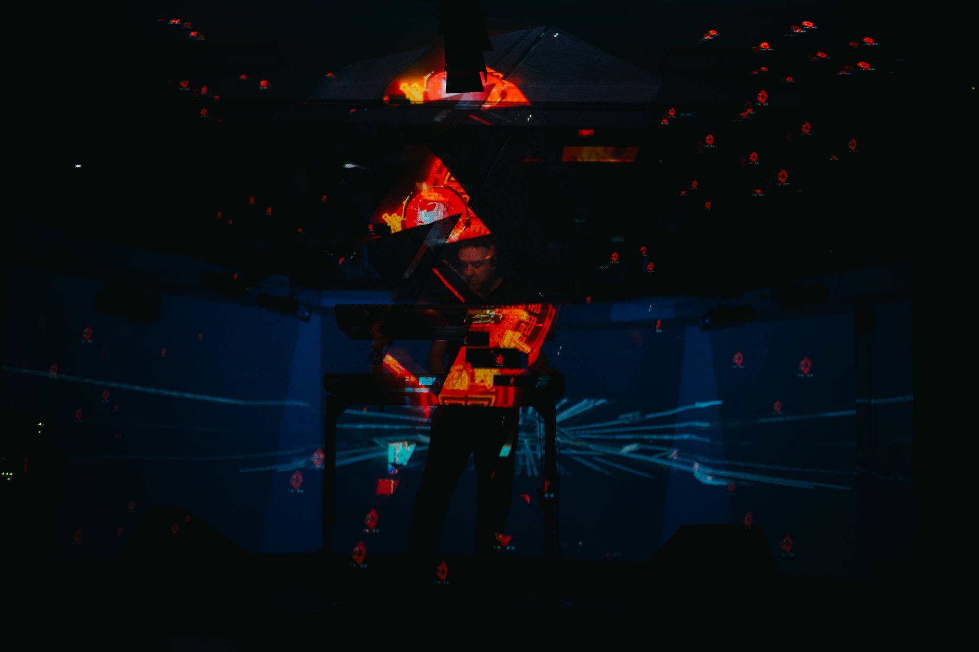 A performer stands at an electronic music setup surrounded by immersive projections of vibrant red and blue digital patterns, creating a futuristic audiovisual environment.