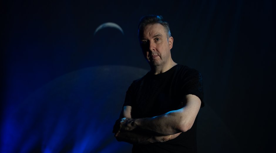 A performer stands with folded arms under dramatic blue stage lighting, with projected moon imagery and beams of light creating an atmospheric backdrop.