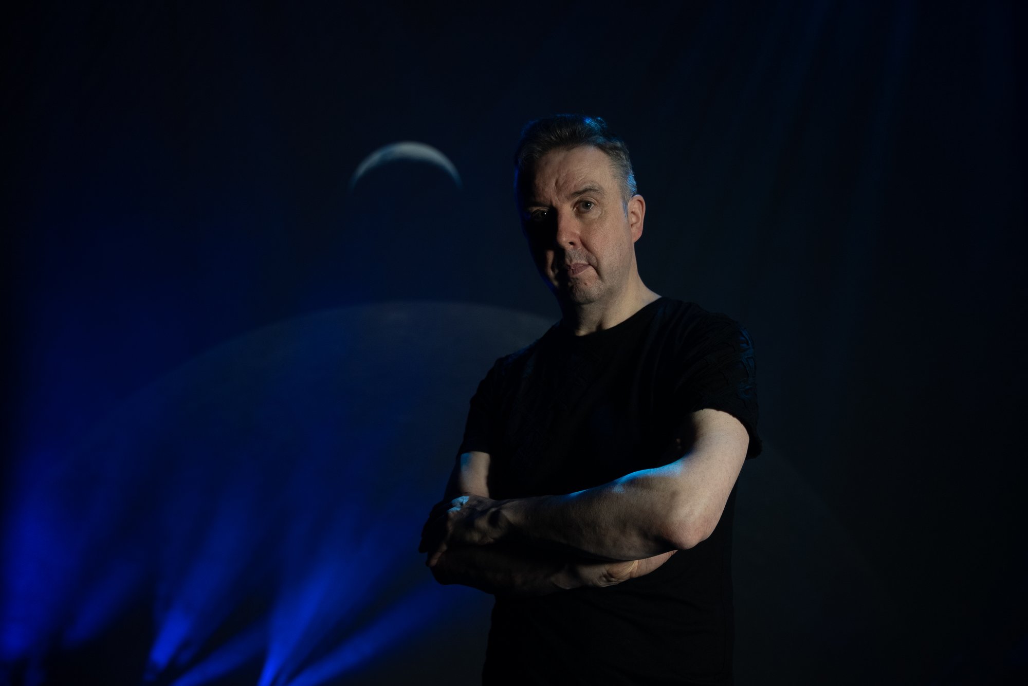 A performer stands with folded arms under dramatic blue stage lighting, with projected moon imagery and beams of light creating an atmospheric backdrop.