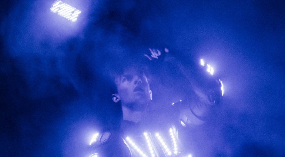 Performer in a glowing LED suit surrounded by blue haze, interacting with illuminated light panels