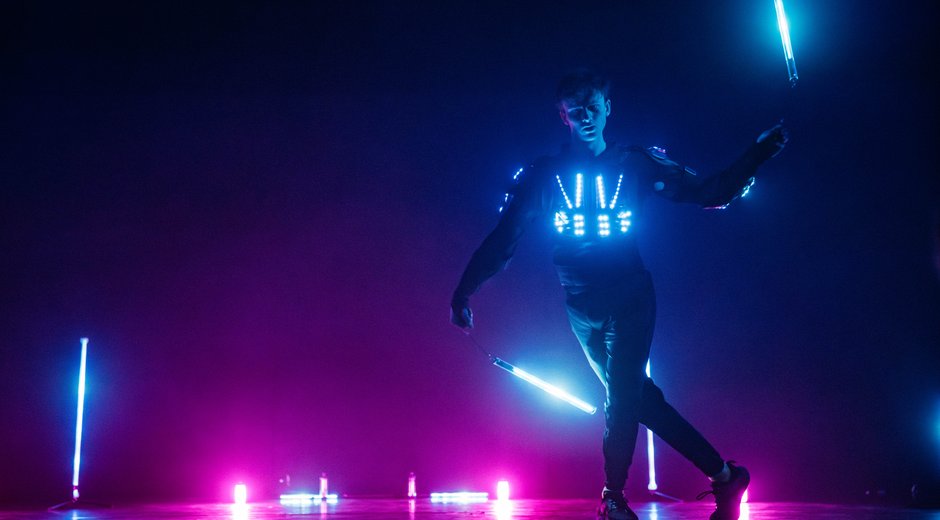 A performer wearing an illuminated LED costume moves with glowing light batons, surrounded by vibrant blue and pink lighting effects.