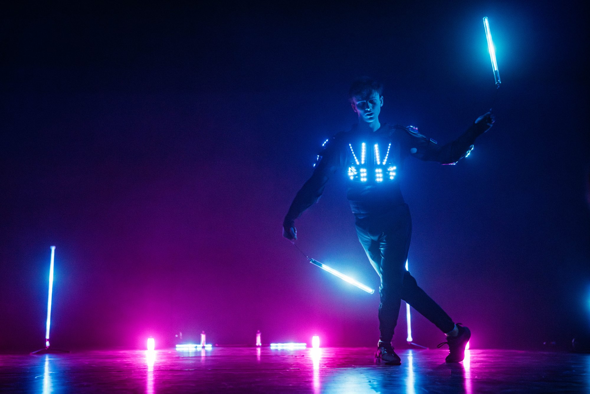 A performer wearing an illuminated LED costume moves with glowing light batons, surrounded by vibrant blue and pink lighting effects.