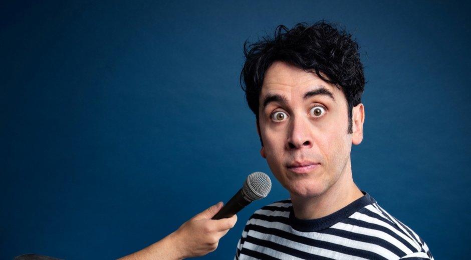 Pete Firman in a striped T-shirt holding a top hat while an arm emerging from the hat holds a microphone toward them.