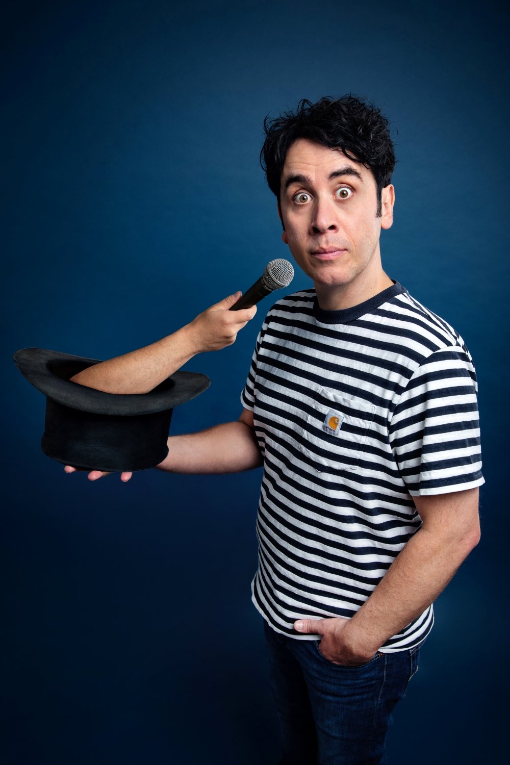 Pete Firman in a striped T-shirt holding a top hat while an arm emerging from the hat holds a microphone toward them.