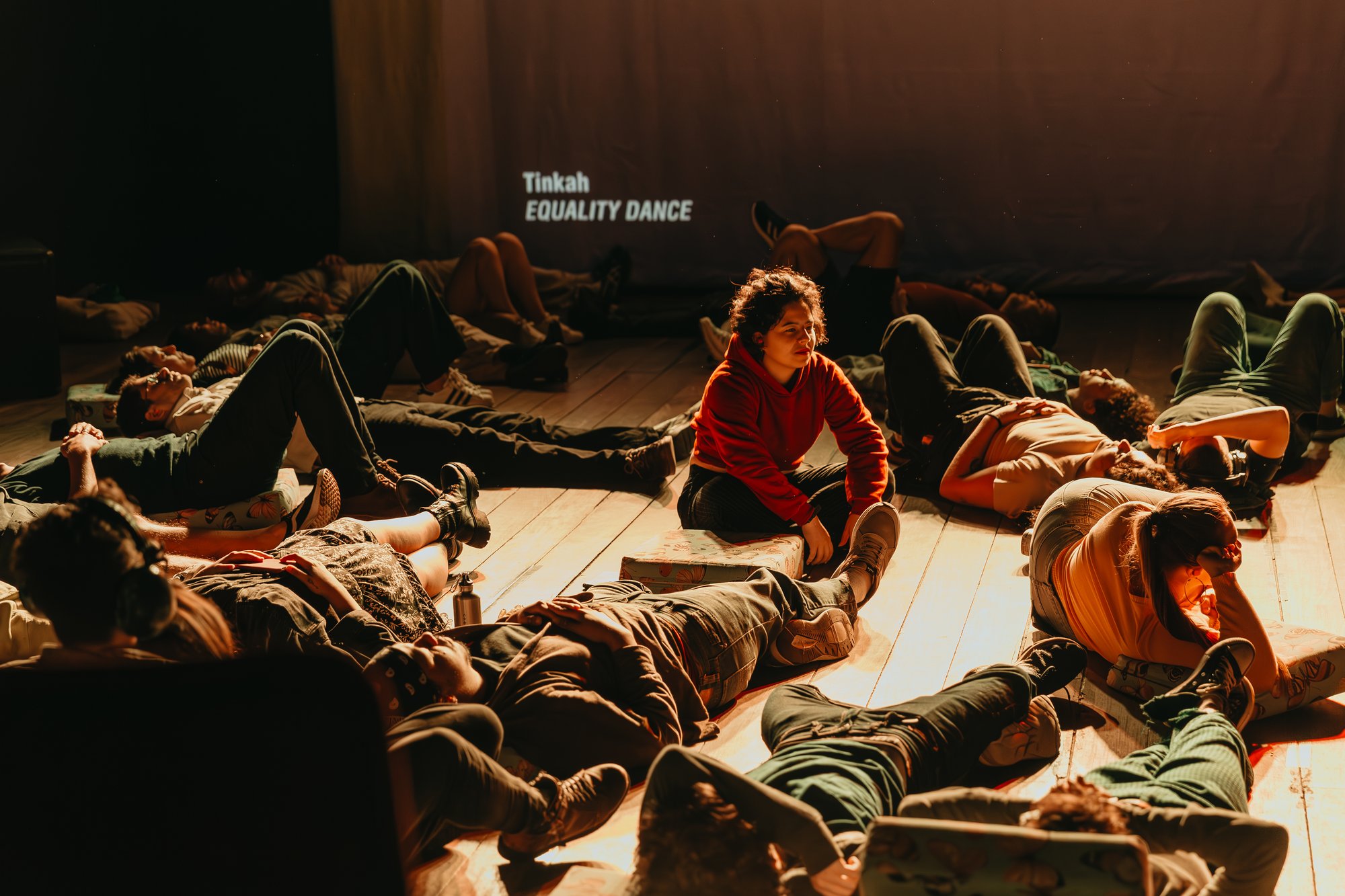 Participants lie and sit across a dimly lit stage during an immersive performance, illuminated by warm amber lighting.