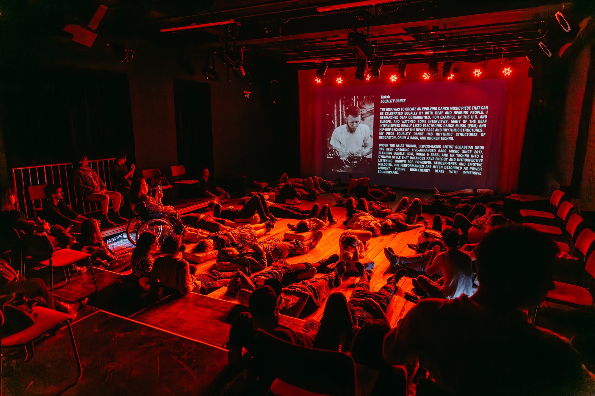 Audience members recline across a performance space bathed in red light, surrounding a large projected screen during an immersive sound experience.