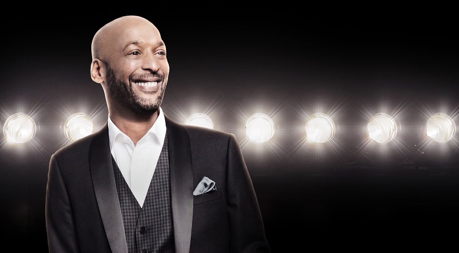 A man wearing a suit in a theatre on a stage with lights with a big smile.