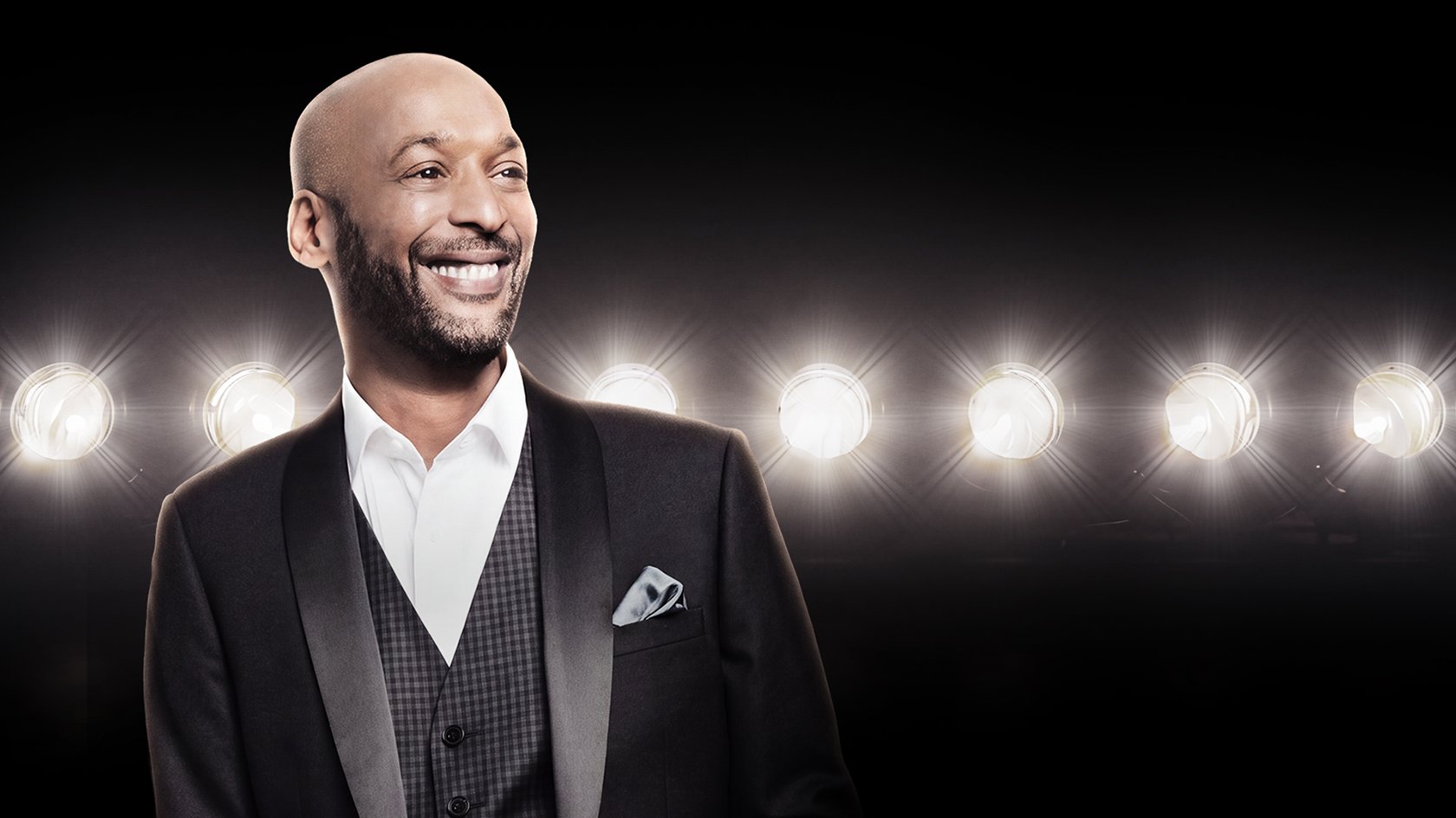 A man wearing a suit in a theatre on a stage with lights with a big smile.