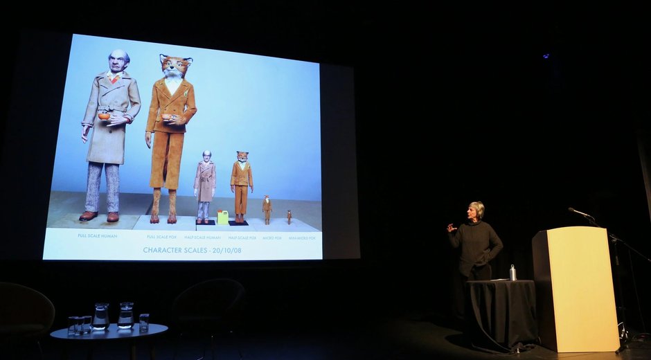 the image captures a speaker presenting character design scale references to an audience in a theatre or conference setting