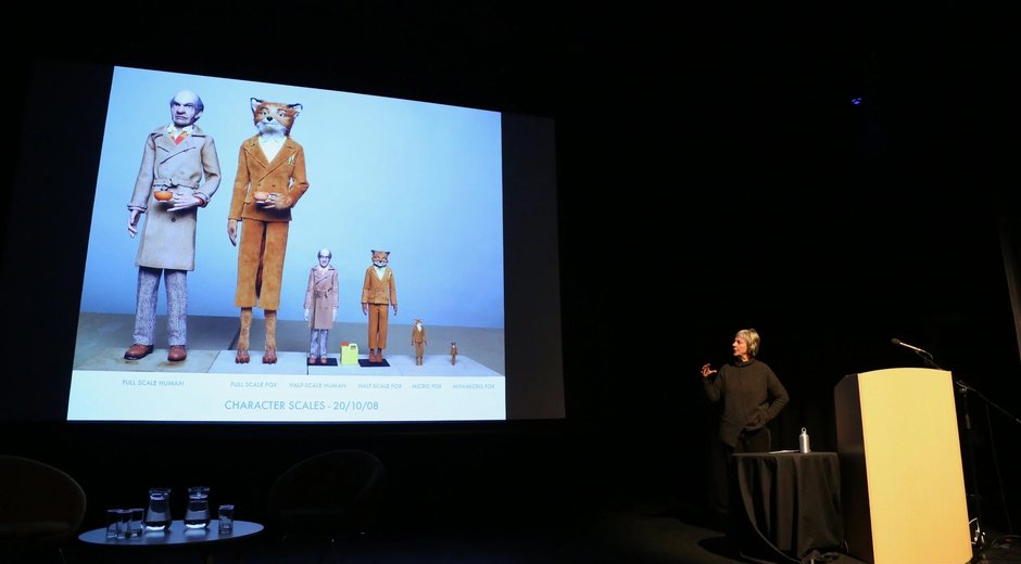 the image captures a speaker presenting character design scale references to an audience in a theatre or conference setting