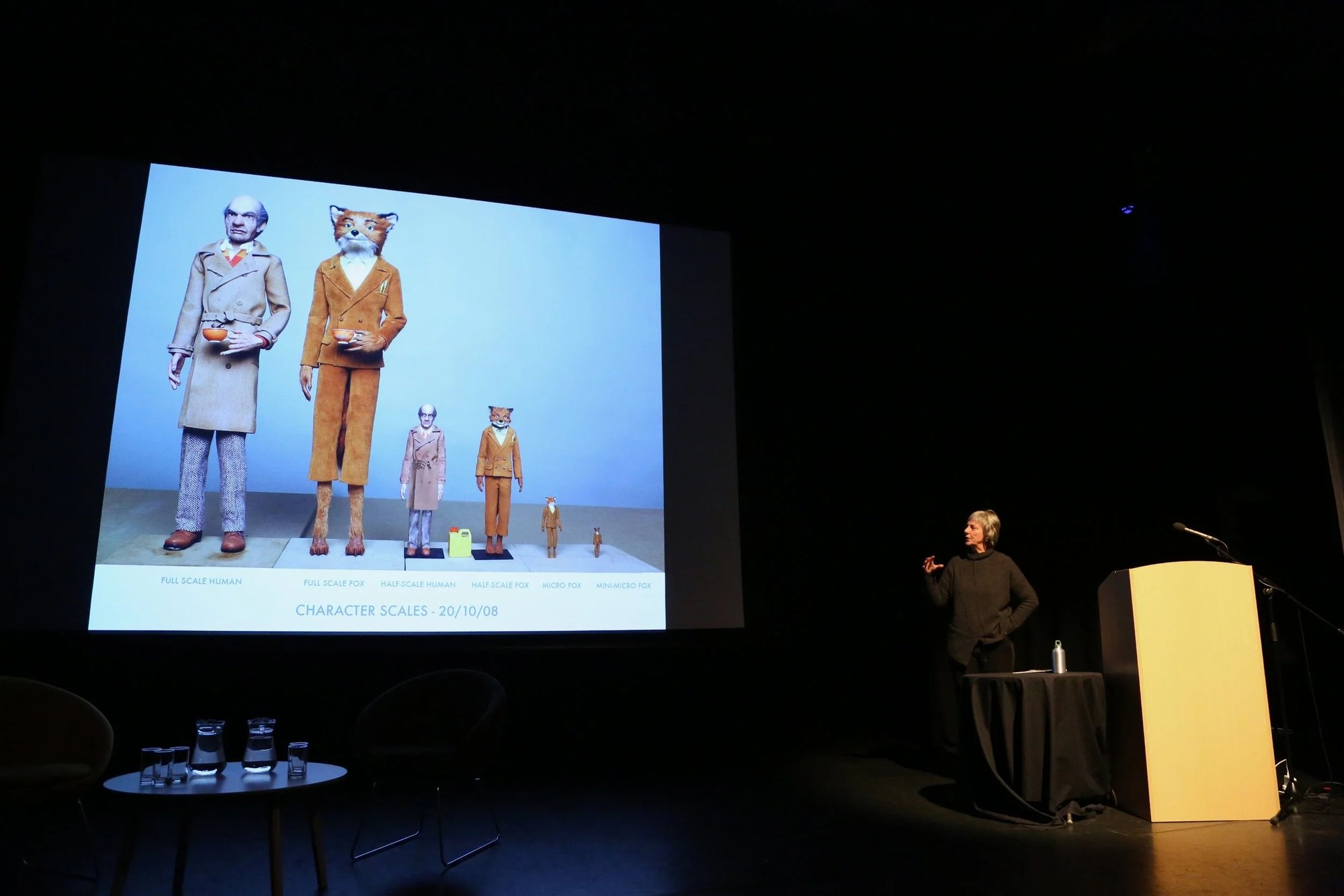 the image captures a speaker presenting character design scale references to an audience in a theatre or conference setting
