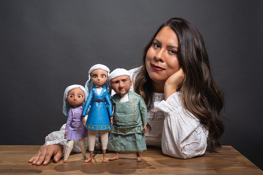 Person in white blouse poses behind three handcrafted puppets on a wooden table