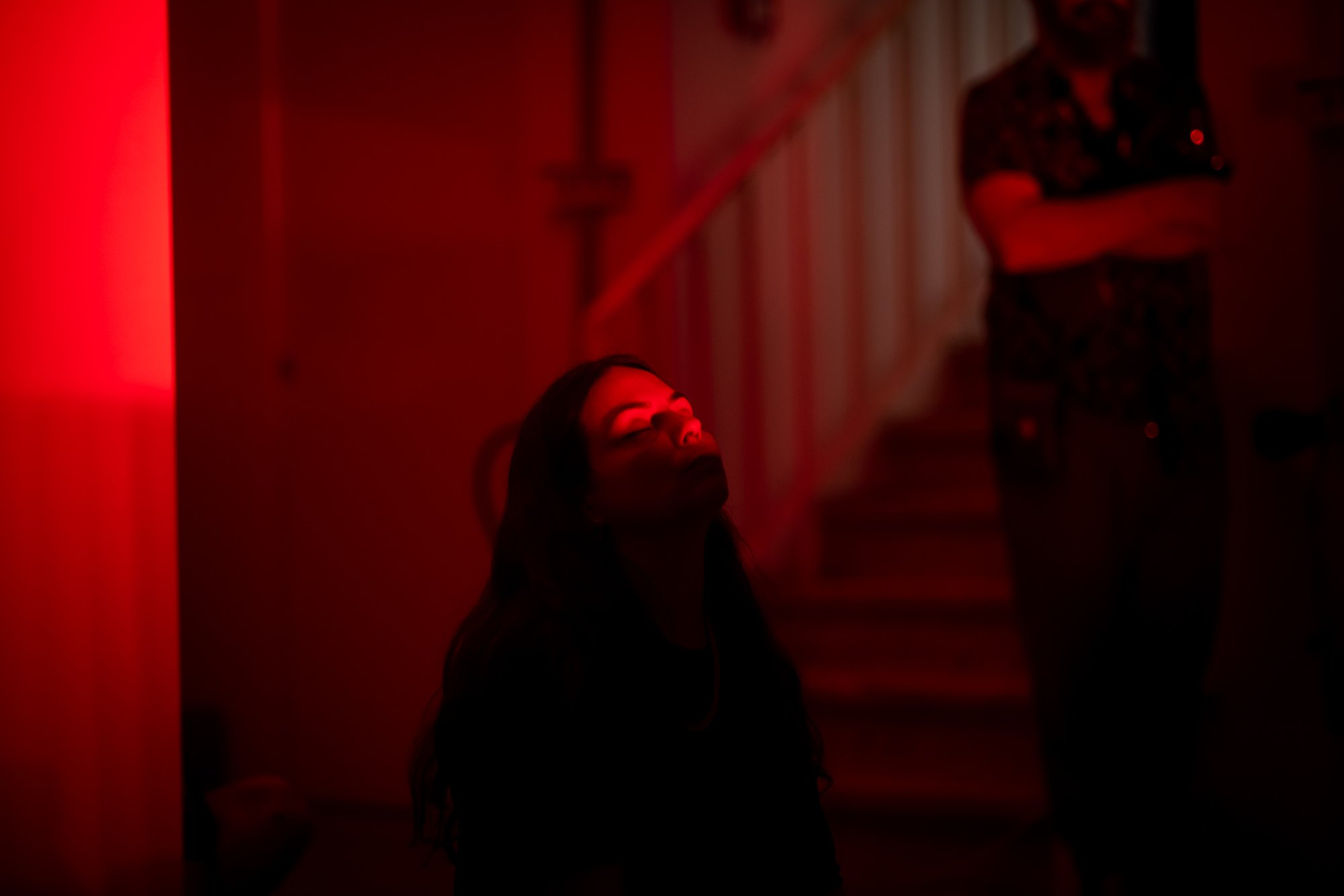 Person seated in a darkened space illuminated by deep red light, with a staircase and other figures blurred in the background.